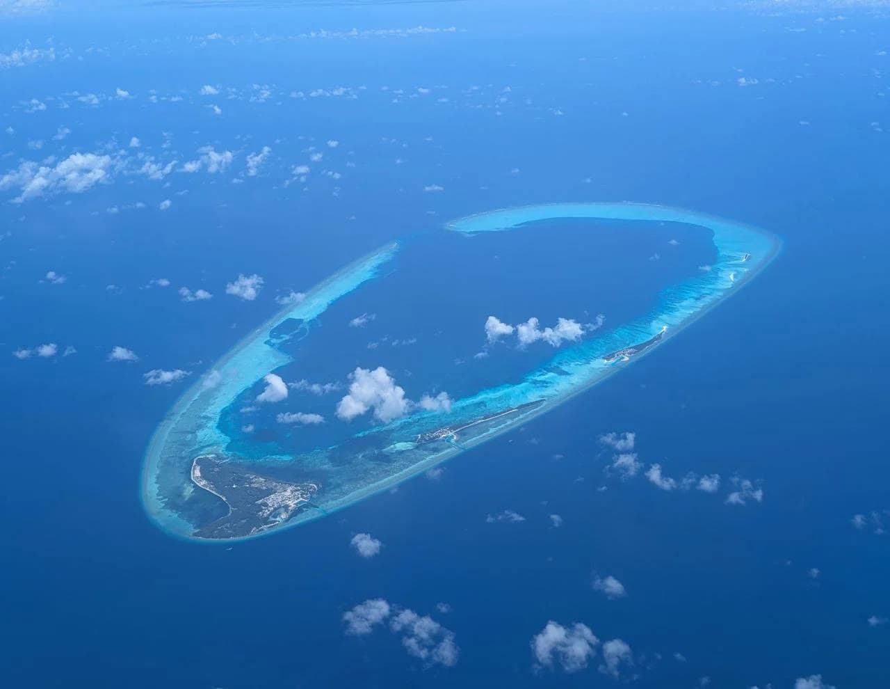 A ring-shaped coral reef in deep blue ocean, clouds throwing shadows across it