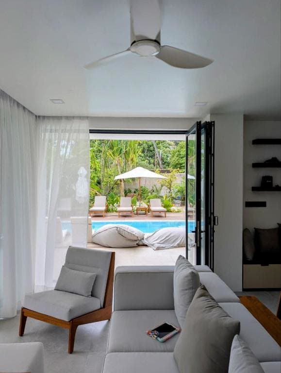 A sofa and armchair beside folding doors opened onto a pool and sun loungers